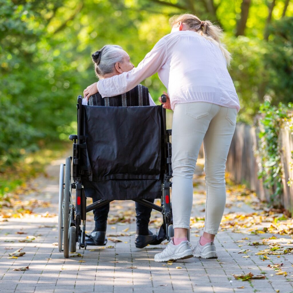 eine junge Frau schiebt eine alte Dame im Rollstuhl durch einen Park