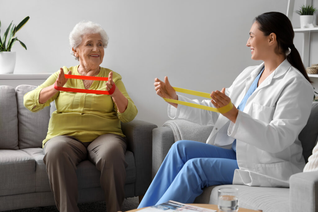 Laechelnde Seniorin und Physiotherapeutin ueben gemeinsam mit Fitnessbaendern